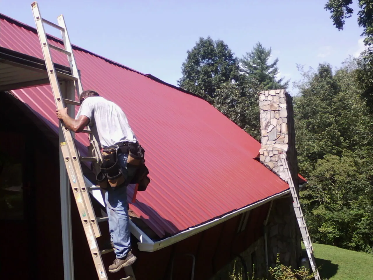 Roofer on a ladder beside a red metal roof and stone chimney — Soffit & Fascia Repair by Roofing Brothers in Ingomar
